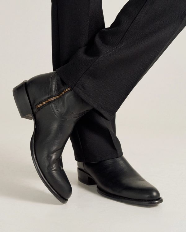 A person wearing black dress pants and black leather boots with a side zipper, standing with one leg crossed over the other.