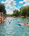 People swimming and lounging on floats in a blue body of water on a sunny day, surrounded by greenery and trees.