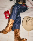 The Annie in Copper Cowhide. Person in fringed leather pants and metallic cowboy boots sitting on a bed, holding a red corded phone; The Annie Cowhide cowboy hat rests nearby.