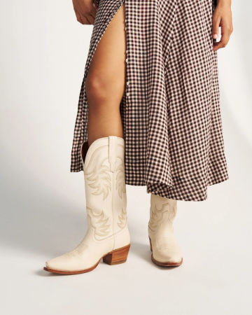 The Annie in Bone Cowhide. Person wearing a brown and white gingham skirt with a high slit and cream-colored bone cowhide cowboy boots, standing against a plain background.