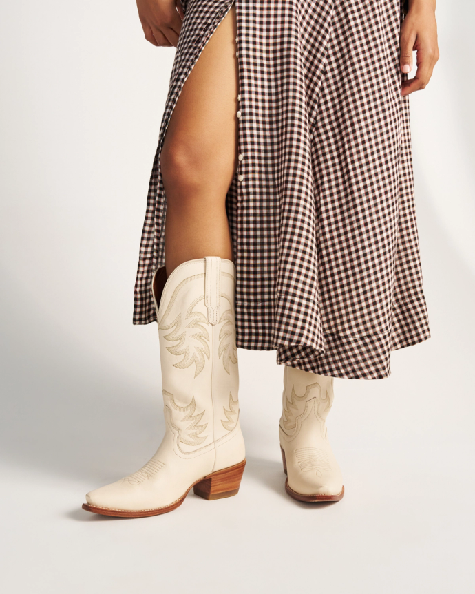 The Annie in Bone Cowhide. Person wearing a brown and white gingham skirt with a high slit and cream-colored bone cowhide cowboy boots, standing against a plain background.