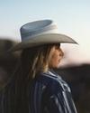 Person wearing a cowboy hat and striped shirt, facing left, with a blurred mountainous background and evening sky.