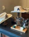 Two hats with feathers displayed on stands next to glass containers of feathers and a wooden tray with two green and two white round pads on a dark countertop.