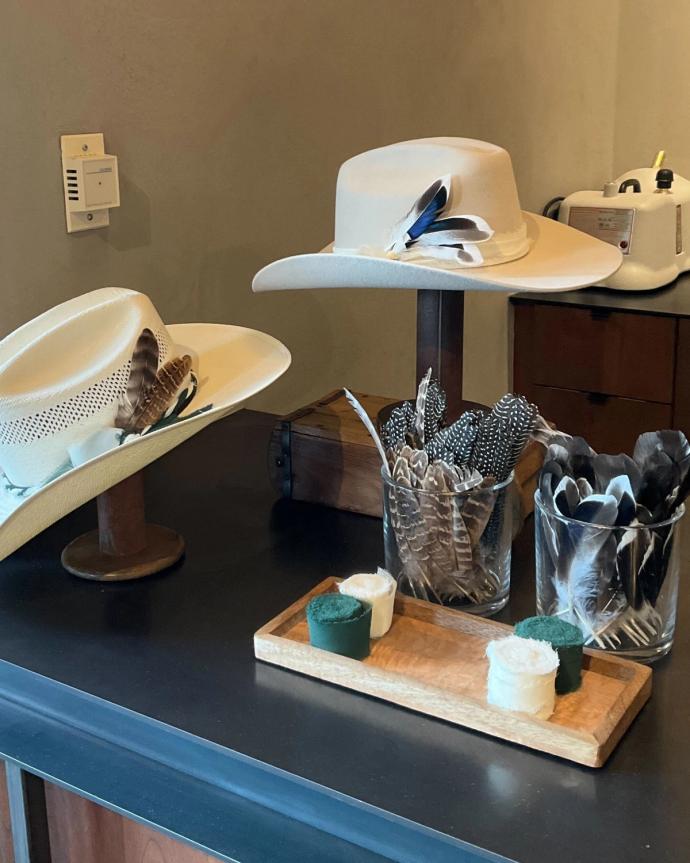 Two hats with feathers displayed on stands next to glass containers of feathers and a wooden tray with two green and two white round pads on a dark countertop.