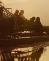Four people ride an airboat through water at sunset, with reflections visible and warm light illuminating the scene.