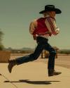 A person wearing a cowboy hat, red jacket, jeans, and boots is mid-jump on a sidewalk in a sunny outdoor setting with mountains in the background.