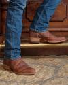 A person wearing blue jeans and brown leather boots stands on a stone floor next to a wooden surface with carved details.