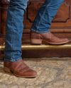 A person wearing blue jeans and brown leather boots stands on a stone floor next to a wooden surface with carved details.
