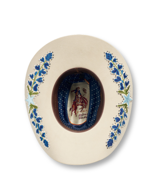 The underside of a cream cowboy hat with blue floral embroidery and a patterned inner band featuring a rodeo figure label.