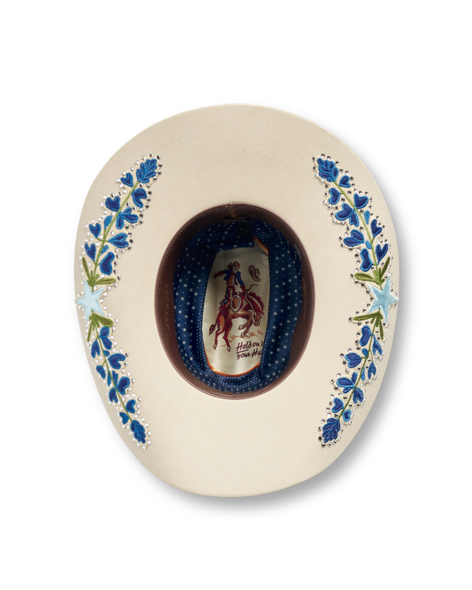 The underside of a cream cowboy hat with blue floral embroidery and a patterned inner band featuring a rodeo figure label.
