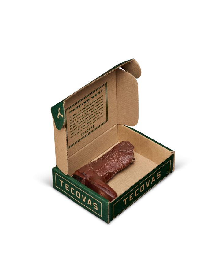 An open Tecovas box displays a small, brown leather item shaped like a cowboy boot, with a printed message on the inside lid.