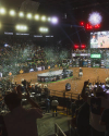 An indoor arena with an audience celebrates as confetti falls onto a central stage. Multiple screens and lights are visible overhead, displaying images and branding. Spectators cheer from the stands.