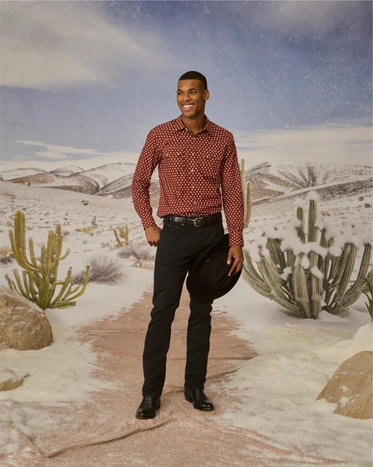 A man, wearing black jeans and a red shirt, standing in a desert with cactus and a cowboy hat.