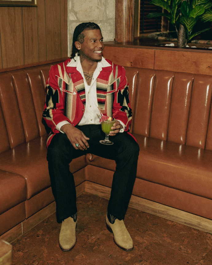 A man sits in a brown leather booth wearing a red and patterned jacket, holding a cocktail with a garnish, smiling and looking to the side.