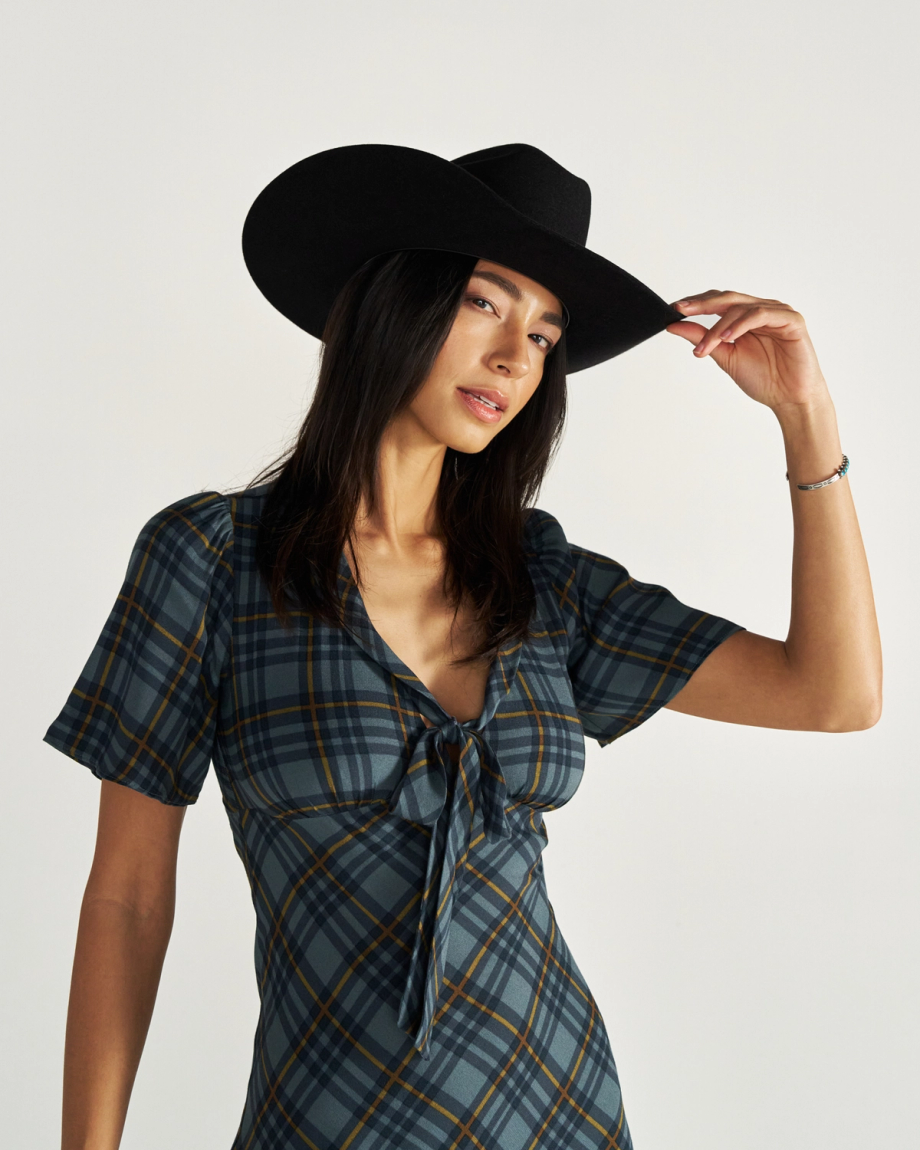 Woman wearing a black cowboy hat and a plaid dress stands against a plain background, holding the brim of her hat with one hand.