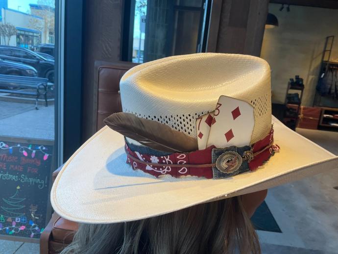 A person wearing a white cowboy hat adorned with a red bandana band, a brown feather, and playing cards, including the nine of diamonds, sits inside an establishment.