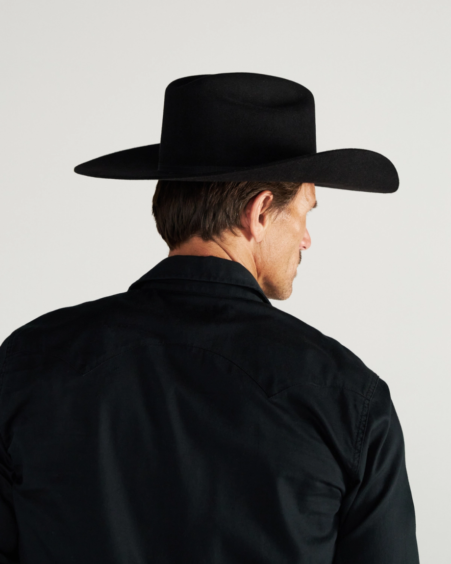Man wearing a black wide-brimmed hat and black shirt, shown from behind against a plain, light background.