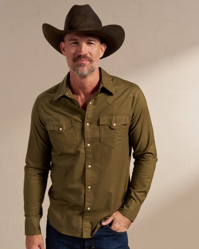 A man with a graying beard wearing a dark cowboy hat and an olive green snap-button shirt stands against a plain background.