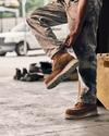 A person rolls up their pant leg while wearing brown work boots and standing on a concrete floor. A pair of shoes is visible in the background.