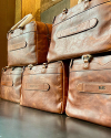 Six brown leather travel bags with embossed name plaques are stacked in two rows on a dark surface against a wooden background.