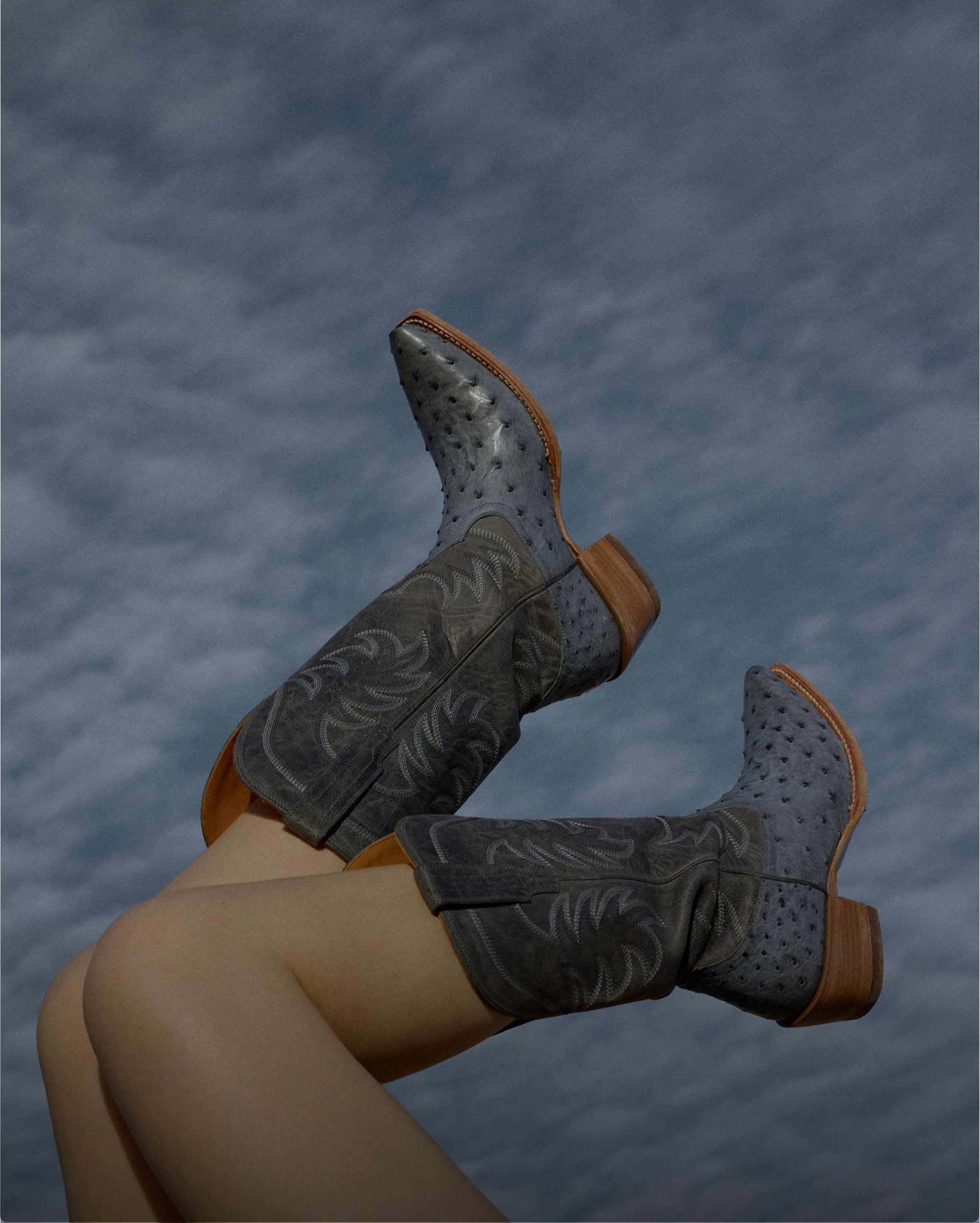 A person with bare legs is wearing dark embroidered cowboy boots, with legs raised against a cloudy sky background.