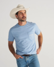 A man in a light blue T-shirt, jeans, and a white cowboy hat stands with one hand on his hip, looking to the side against a plain background.