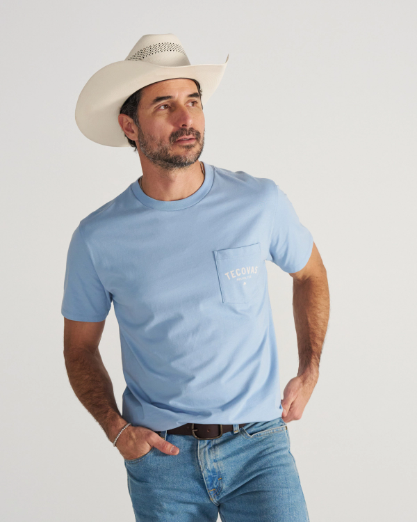 A man in a light blue T-shirt, jeans, and a white cowboy hat stands with one hand on his hip, looking to the side against a plain background.