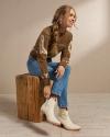 Woman sitting on a wooden stool in a photo studio wearing jeans, a sweater, and white ankle boots