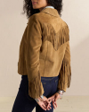 A person is shown from the back wearing a tan suede jacket with fringe detailing on the back and sleeves. They have long dark hair and are holding their hands behind their back.