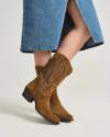 A person wearing a blue denim skirt and brown suede cowboy boots stands against a plain background.