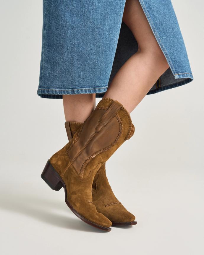 A person wearing a blue denim skirt and brown suede cowboy boots stands against a plain background.