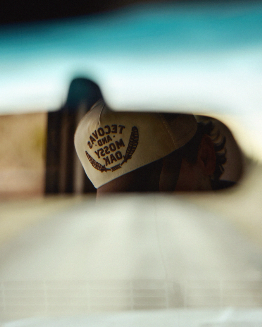 A person wearing a cap that reads "Taco Day and Mussy Oak" is partially visible in a car’s rearview mirror.