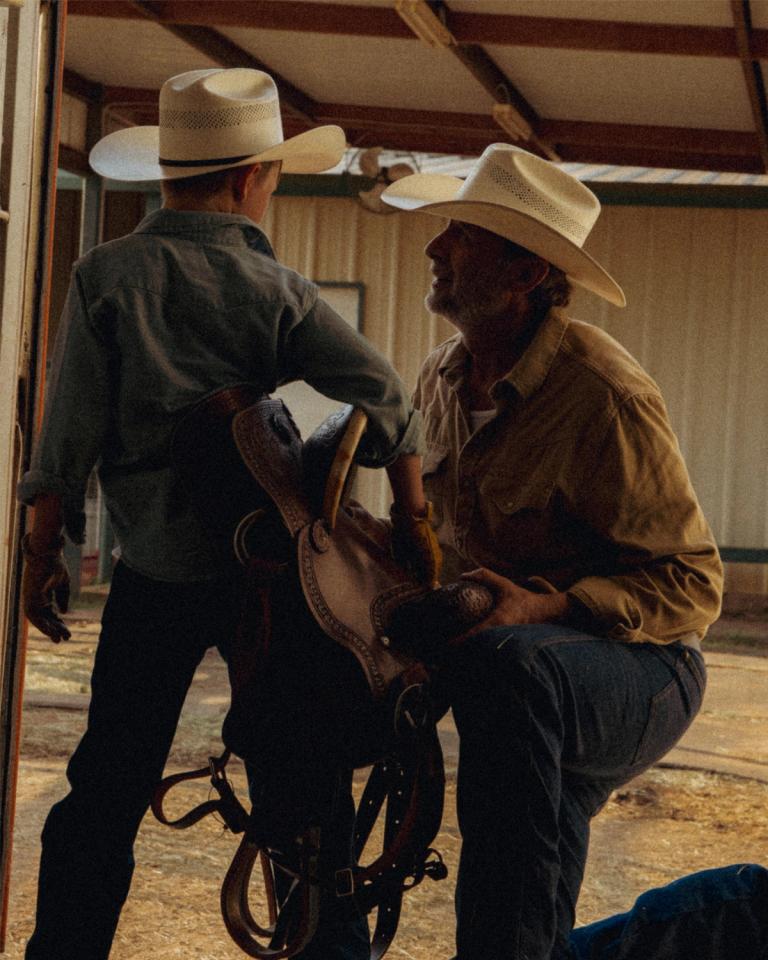 An adult and a child wearing cowboy hats stand together in a barn doorway, holding a horse saddle.