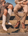 Two people sitting on a fence wearing brown pants and tan cowboy boots, next to a person in a floral dress and white cowboy boots, set against a grassy landscape.