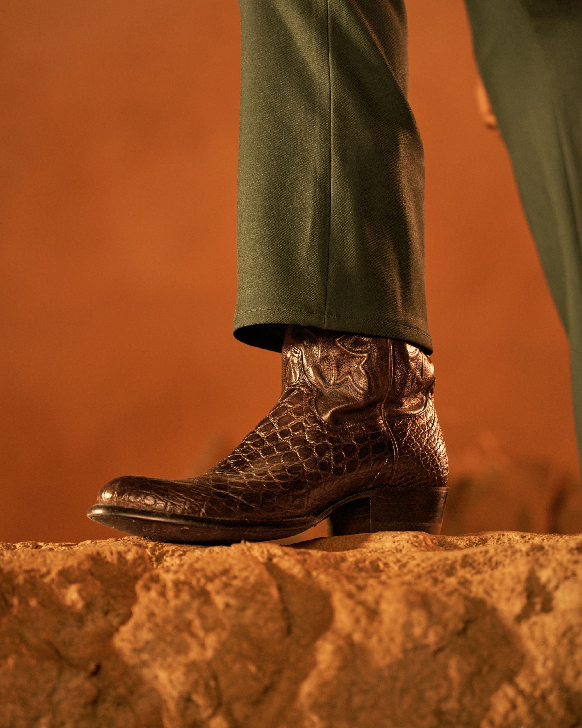 A close-up of a person wearing a black, textured cowboy boot with a pointed toe, standing on a sandy surface.