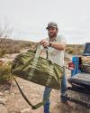 A bearded man in a cap and white shirt lifts a large green duffel bag into a blue pickup truck in a rugged outdoor setting.