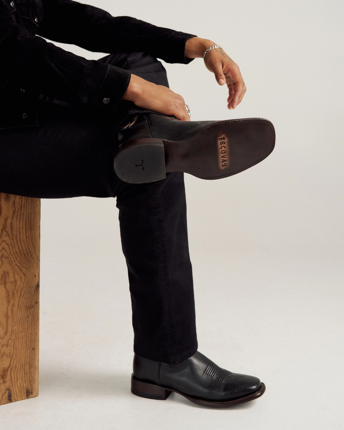 A person in black pants and boots sits on a wooden bench with one leg crossed, showing the sole of the boot stamped with the word “TEXAS.”.