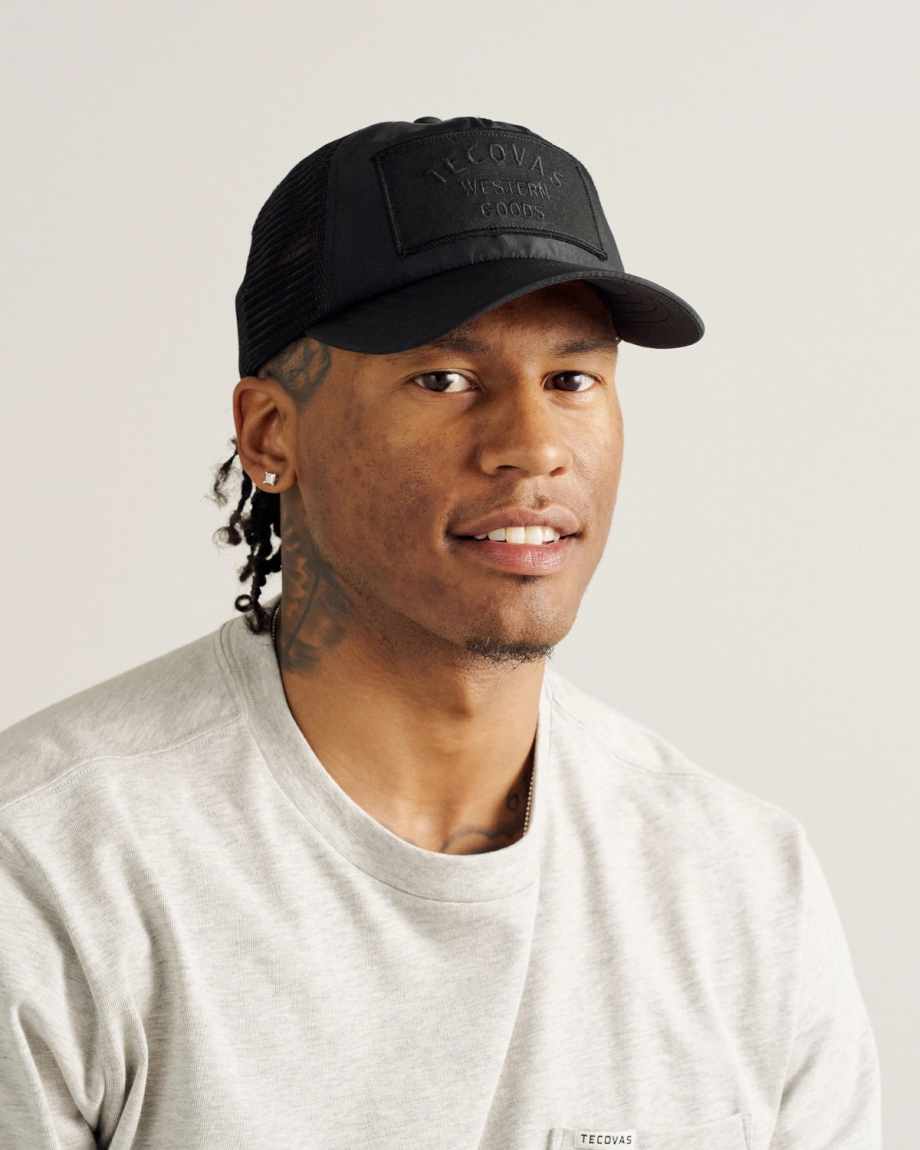 A man wearing a black Tecovas cap, a light gray shirt, and earrings, with visible tattoos on his neck, is sitting and looking at the camera against a plain background.