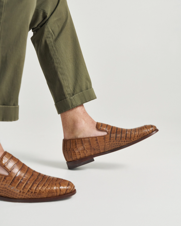 The Bennett in Saddle Alligator. A person wearing olive green pants and brown saddle alligator loafers, with one foot slightly raised against a plain light background.