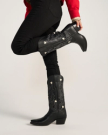 The Annie in Midnight Cowhide. Person wearing Midnight Cowhide embroidered cowboy boots with white heart shapes, black pants, and a red jacket, posing with one leg bent at the knee.