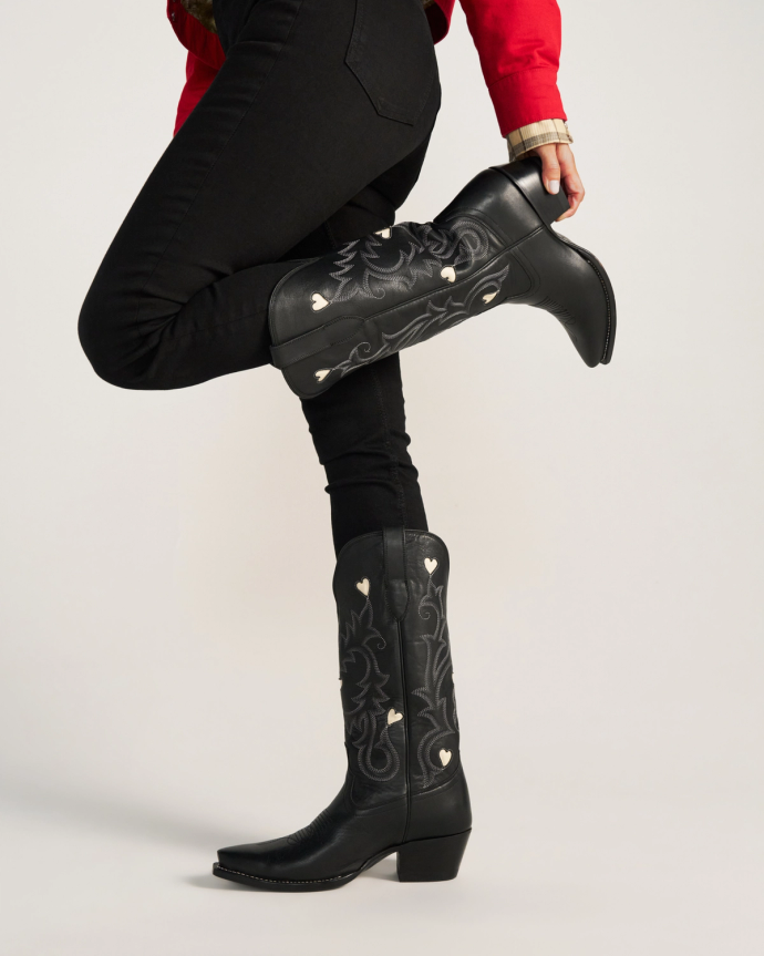 The Annie in Midnight Cowhide. Person wearing Midnight Cowhide embroidered cowboy boots with white heart shapes, black pants, and a red jacket, posing with one leg bent at the knee.