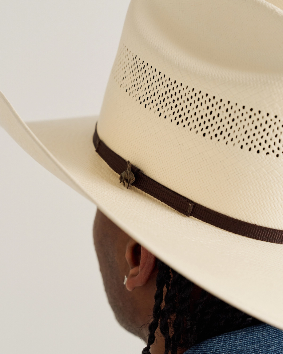 A person wearing a light-colored, wide-brimmed cowboy hat with a brown band and perforated design, shown from the back.