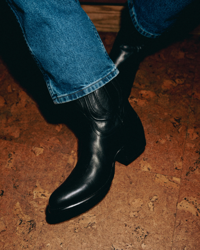 Person wearing black cowboy boots with blue jeans on a brown floor.