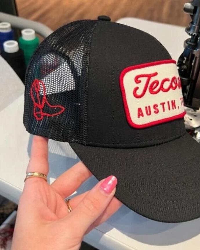 A hand holding a black mesh trucker hat with a red and white patch that reads "Tecovas AUSTIN, TX" and a red embroidered cowboy boot on the side.