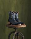 A pair of black cowboy boots with floral embroidery sits on a wooden stool against a green backdrop.