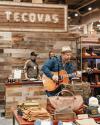 A musician plays an acoustic guitar at a Tecovas booth with various leather goods and clothing displayed on tables. Two people are in the background near a wooden wall with the Tecovas sign.