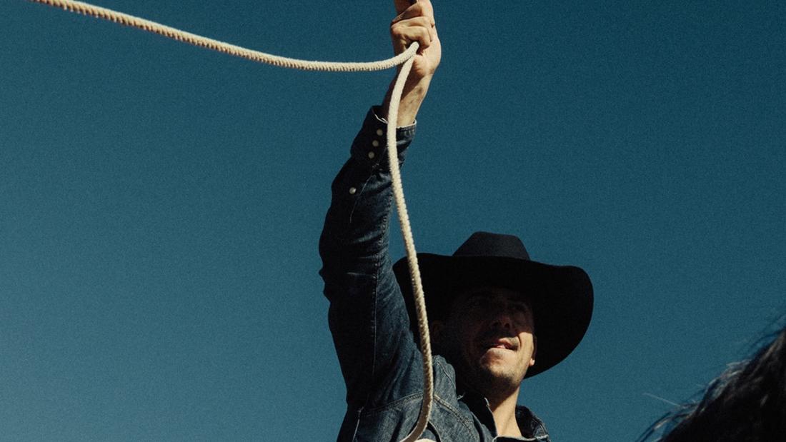 White cowboy hat with decorative engravings, including mountains, a sun, a moon, and cacti, placed on a wooden surface with an abstract painting in the background.