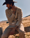 Man wearing a cowboy hat and beige sweater sits on a wooden fence in a rural field.