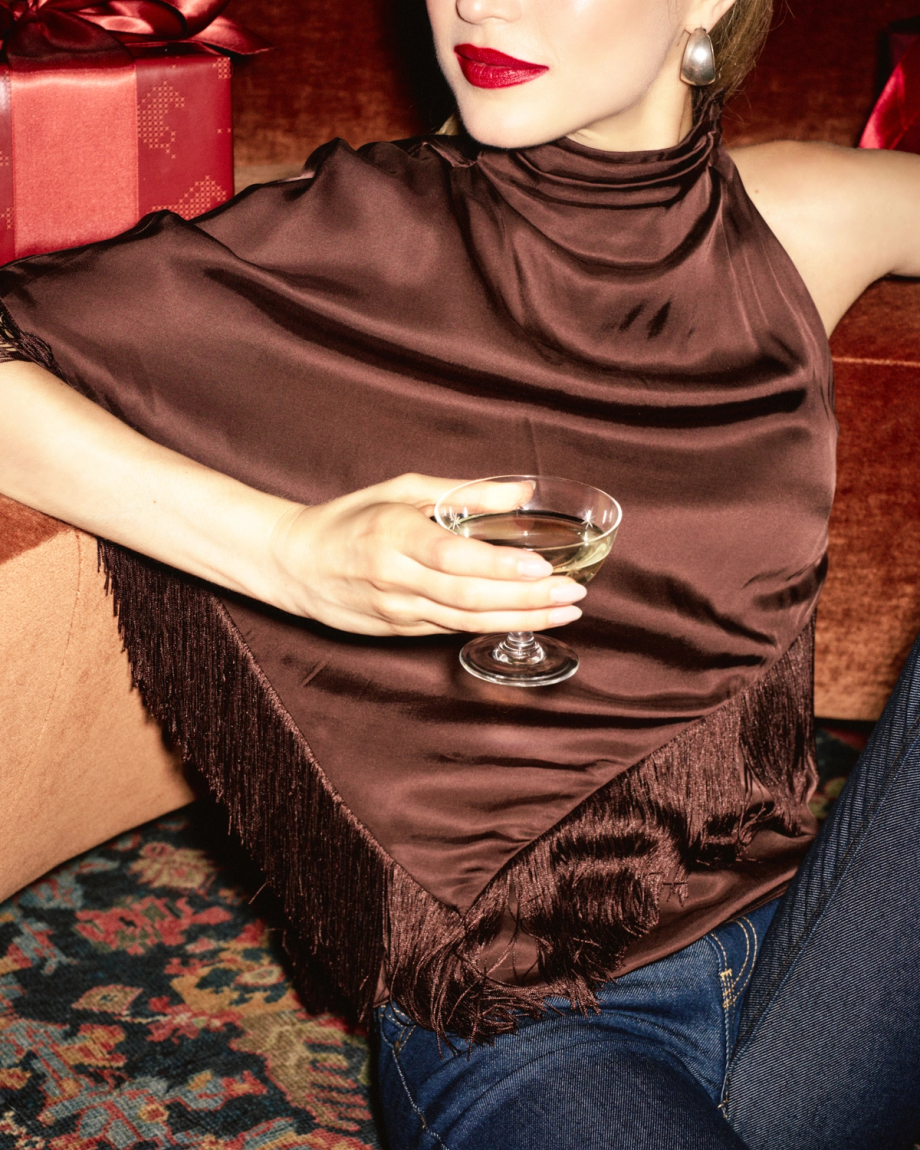 A person wearing a brown high-neck top with fringe trim holds a glass of wine, sitting next to a red gift box.