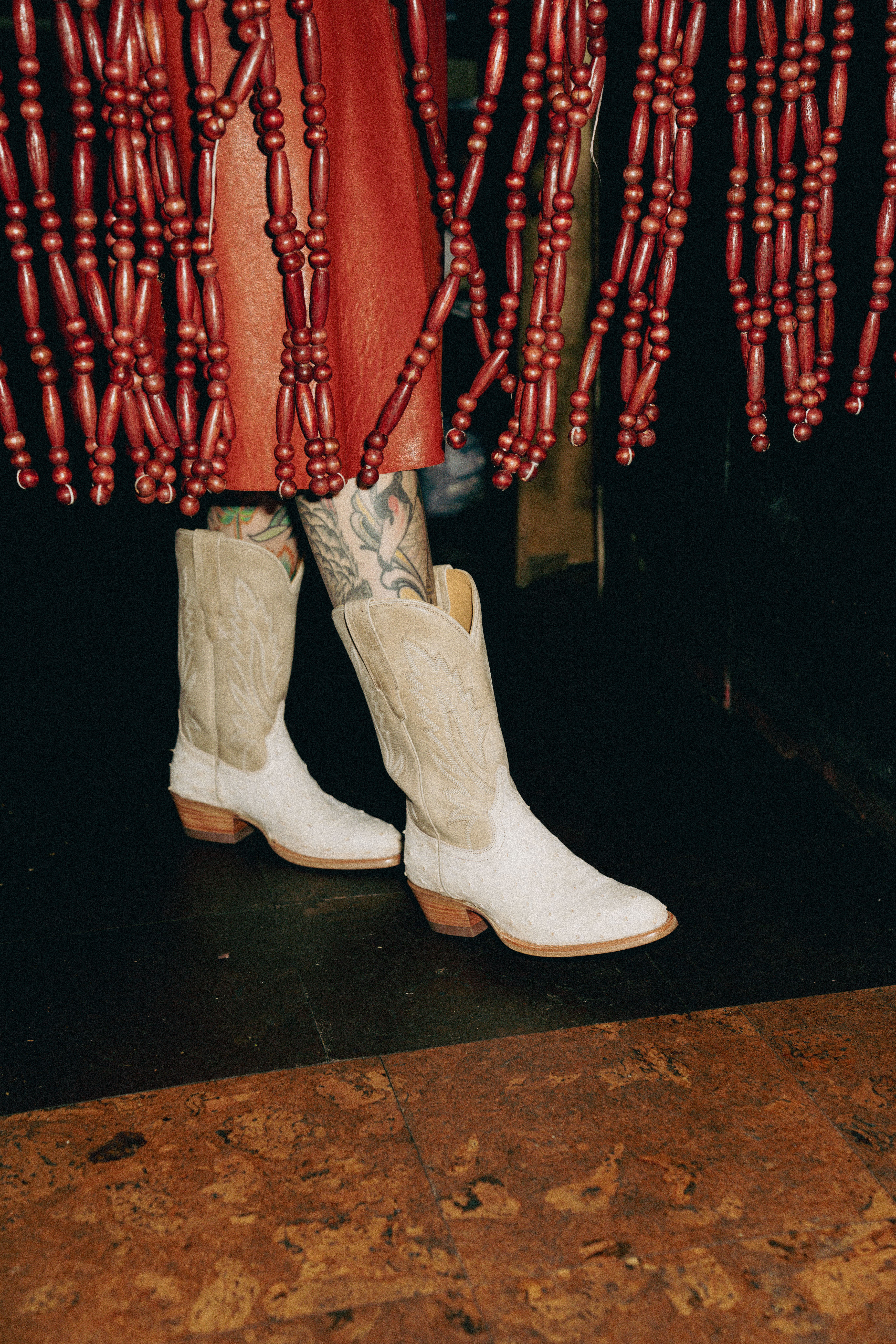 Person in white cowboy boots and tattooed legs under a red skirt stands next to a red beaded curtain on a wooden floor.